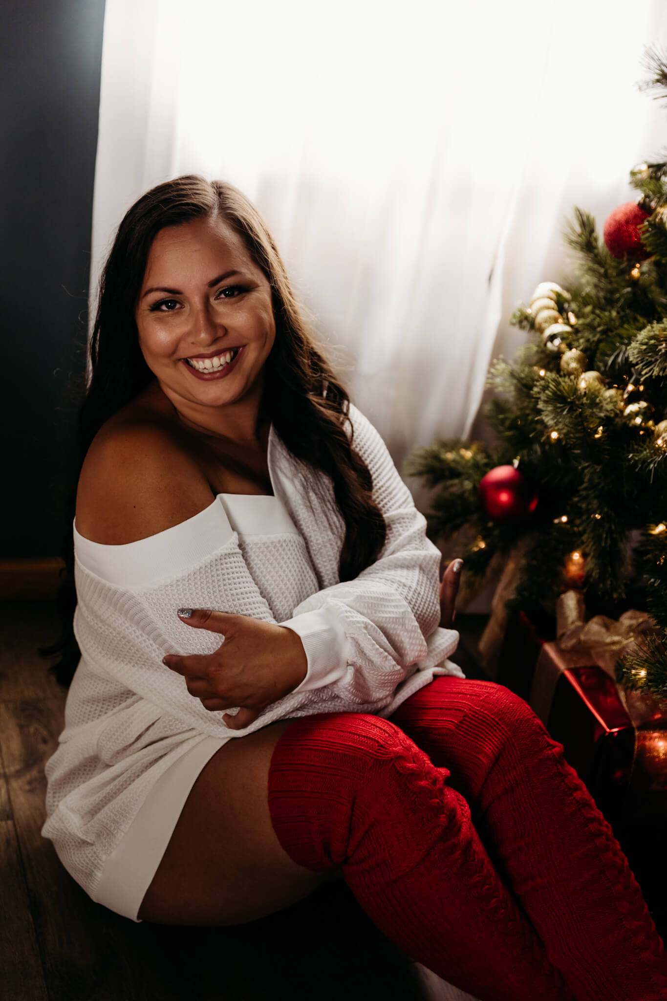 woman in a white sweater and red tall socks hugging herself while sitting by the Christmas tree during Tiara's experience
