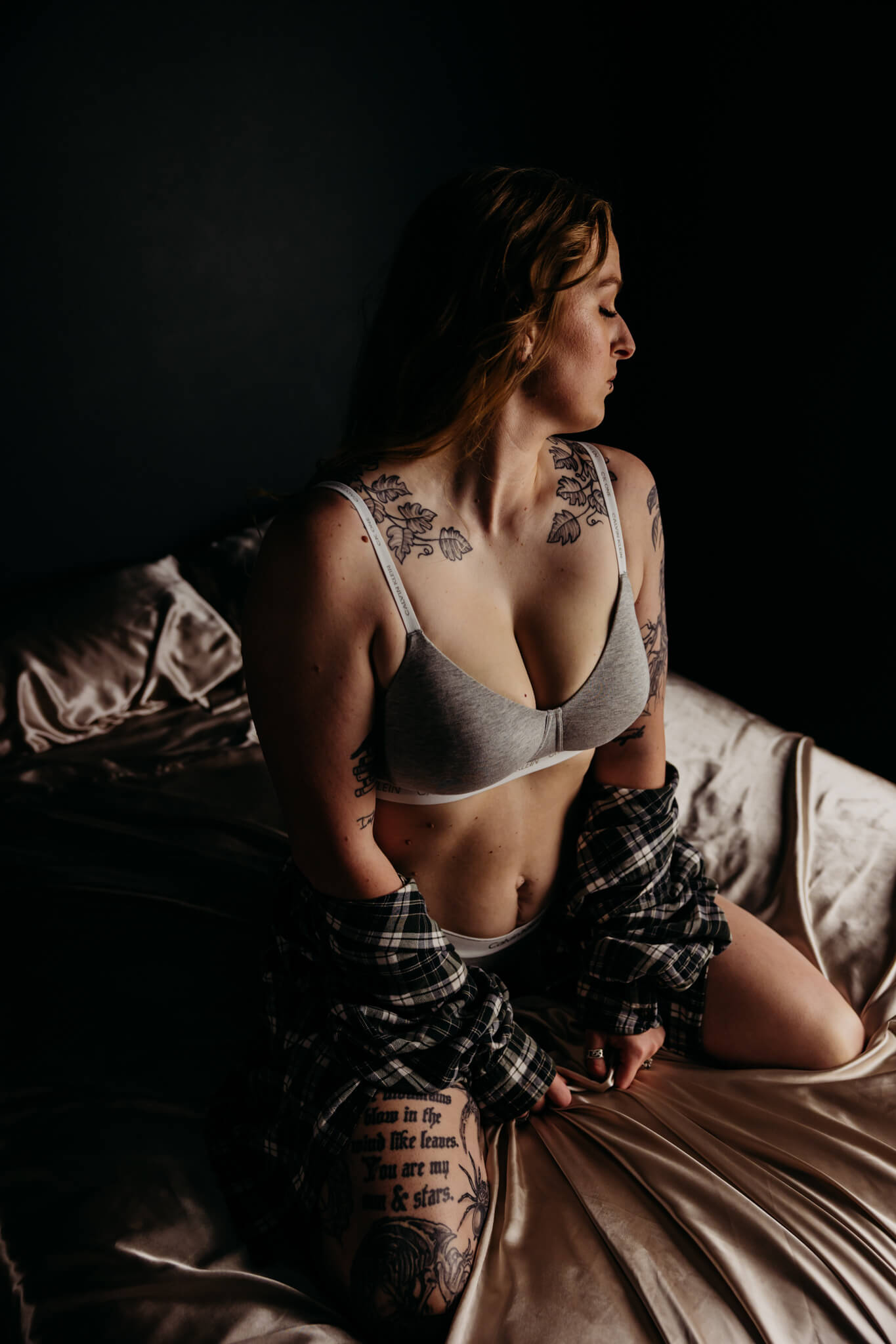 woman kneeling on a bed as she drapes a flannel over her calvin klein undies and looks away